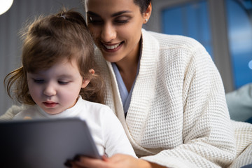 Smiling lady and her little daughter holding gadget at home