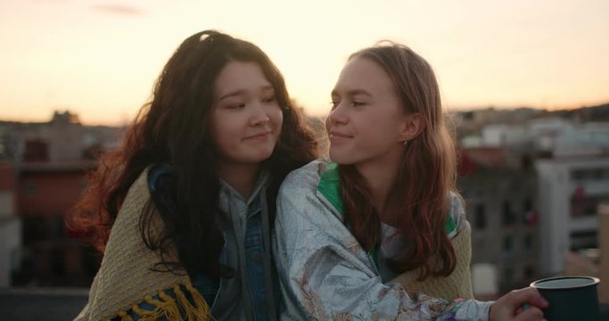Portrait of two cute young female friends sharing blanket at windy rooftop outdoor, attractive mixed race girls smiling looking at each other enjoying hot drink from cup friendship and youth concept