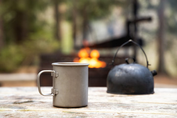 Cup of coffee and camp kettle on the wooden table. Camping concept. Campfire in forest.