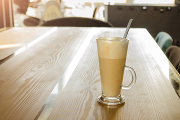 Tall transparent glass beaker with a handle, with foam latte inside and spoon, on a wooden table in a cafe in the sunshine