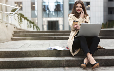 young beautiful businesswoman having phone conversation works in her laptop drinks coffee sitting...