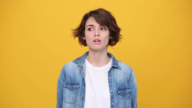 Boring cute brunette young woman in denim jacket white t-shirt posing isolated on yellow background in studio. People lifestyle concept. Looks around, bites her lips, misses and waiting for something