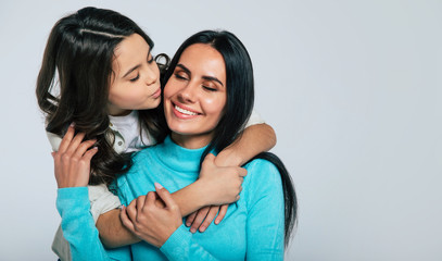Let me kiss you, mom. Charming woman with long dark hair is smiling and closing her eyes while her little child is hugging her from the back and kissing in the cheek.