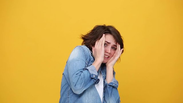 Scared shocked brunette young woman in denim jacket white t-shirt posing isolated on yellow background in studio. People sincere emotions, lifestyle concept. Looking at camera covering hiding face