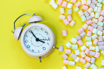 Top View of Pastel Shaped Marshmallow Candies and alarm clock with Some Scattered on the Pale yellowTable
