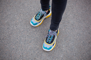 Feet shod in sneakers multi-colored yellow, white, black and blue are on the asphalt road. Close-up