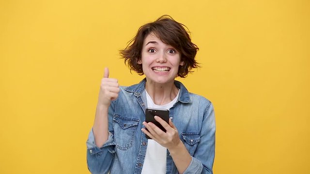 Smiling excited young woman in denim jacket white t-shirt posing isolated on yellow background in studio. People sincere emotions, lifestyle concept. Pointing on cellphone using mobile phone happy win