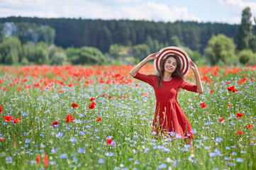 Dreamy woman in red dress and a big red striped hat in beautiful herb flowering poppy field....