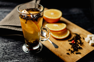  Citrus drink, tea with petals in a transparent glass mug with slices of sweet orange and refined sugar. Slicing fruit on a wooden Board with a brown napkin in a cozy dark background in a low key