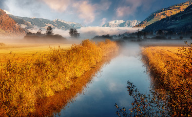 Wonderful picturesque Scene. Awesome Autumn landscape with colorful sky, reflected on the calm river. Amazing nature counryside. Impressive Misty Morning. Fantastic alpine scenery