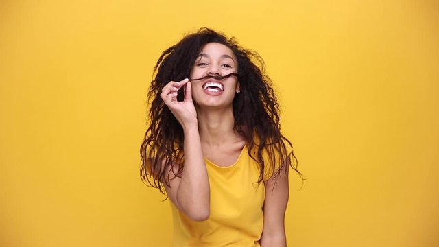 Young african american girl in yellow t-shirt posing isolated on orange background in studio. People lifestyle concept. Looking at camera charming smile play with curly hair making mustache under nose