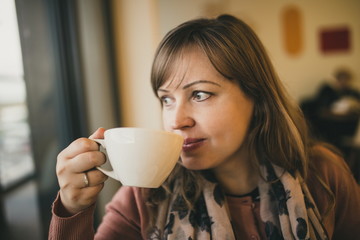 Beautiful young woman drinking coffee sitting by the window in the house. Hot coffee. Beautiful young girl drinking tea or coffee in café. Beauty model woman with the cup of hot beverage. Warm filter