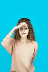 Portrait of a young beautiful woman wearing sweatshirt makes a visor with palm over the eyes and looking at you coquettishly isolated over blue background