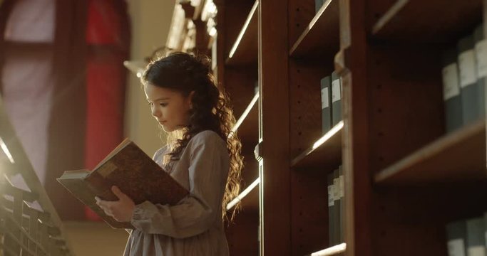 Small brunette Caucasian beautiful and cheerful girl is excited with interesting story. Little pretty schoolgirl standing in dark magical bibliotheca and reading book. Female teen reader in library.