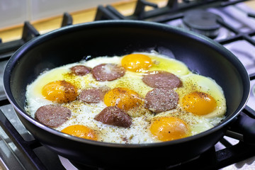 Fried eggs with sausage and seasonings in a black pan on a gas stove. Tasty and nutritious breakfast for the family. Selective focus. Closeup view