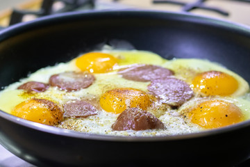 Fried eggs with sausage and seasonings in a black pan on a gas stove. Tasty and nutritious breakfast for the family. Selective focus. Closeup view