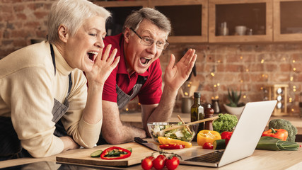 Elderly Couple Looking At Laptop Screen In Kitchen And Emotionally Gesturing