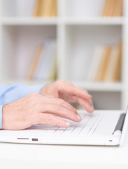 Hands of a modern elderly man lying on the keyboard of a white laptop on the background of shelving with books