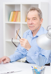 An elderly man sitting near a laptop, holding glasses near his face and looking to the camera