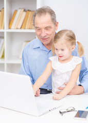 Fototapeta premium An elderly man and a little girl sitting on his lap sitting in a room in front of a laptop