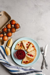 Pancakes with raspberry jam on a pan next to chicken eggs whisk and lemon on a plate on a light concrete background. The view from the top.  side view. space for text