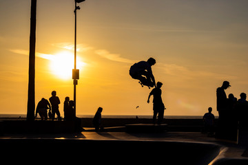 Horizontal image of silhouettes and shadows of skaters at sunset in the skateboard park of Venice Beach in California, figure in the air, Pacific ocean in background