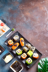 Sushi rolls set served on black stone slate by sauce and nori sheets on dark background with evergreen leaf.
