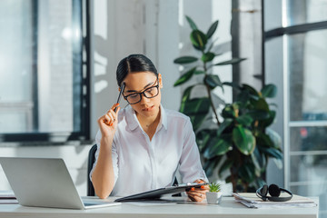 pensive asian translator working with laptop and documents in modern office