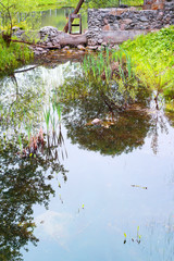 Stone bridge with old dam through the narrow river, clearly reflections of trees and bushes on the surface of water.