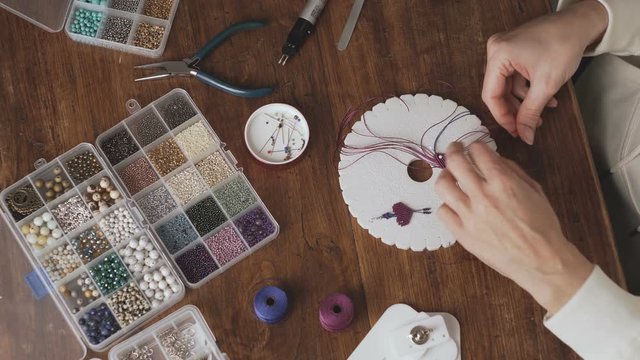 Lifestyle concept, work from home to reinvent your life: view from top of woman hands making macrame knotted jewelry with stone beads and tools on wooden table