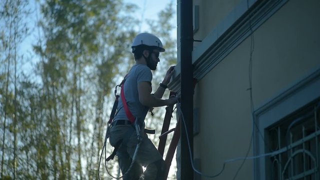 Young caucasian male apprentice industrial worker/repairman climbing up ladder leaning against house wall wearing protective white hardhat and safety equipment, safety first at work icon