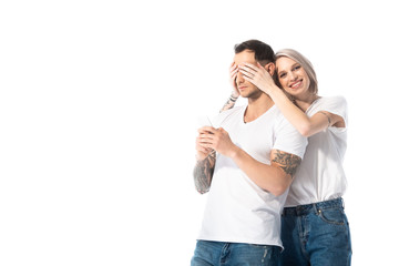 smiling young tattooed girl covering boyfriend eyes while man using smartphone isolated on white