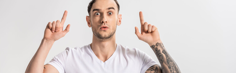 shocked handsome young tattooed man pointing with fingers up isolated on white, panoramic shot