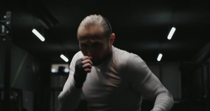 Strong adult man doing shadow boxing and performing various punches during workout in dark gym.