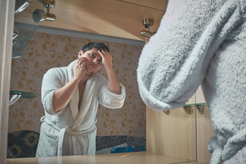 A young man, a Caucasian, stands near a mirror, combing his hair in a white bathrobe in a home appliance © Duha