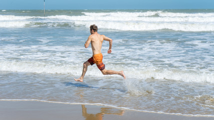 Handsome man with athletic body running around the beach line near the sea.Fit runner on the beach with sea waves background. Beach activities concept.