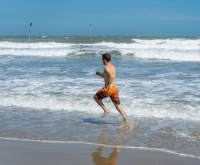 Handsome man with athletic body running around the beach line near the sea.Fit runner on the beach with sea waves background. Beach activities concept.