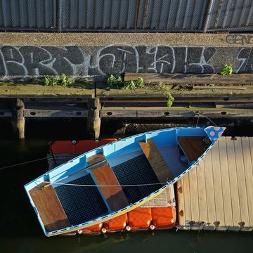 View from overhead of a blue rowboat on a red mat next to a yellow dock next to a wall with graffiti.