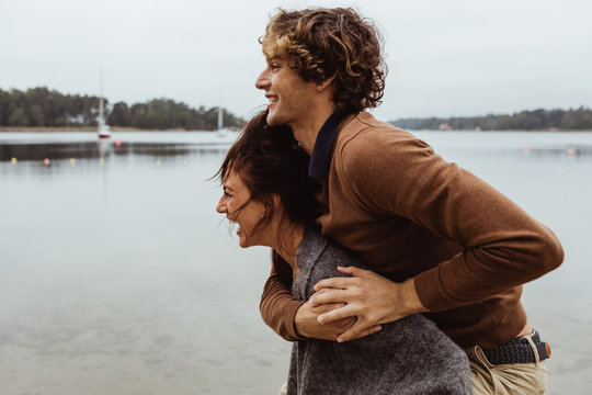 Smiling woman giving piggyback ride to male friend by sea against sky