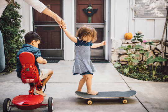Daughter skateboarding with help of mother while son sitting on tricycle