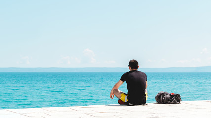 Fototapeta na wymiar young man traveler alone with backpack near him meditating looking into the ocean with copyspace