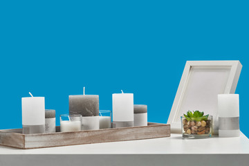White table with green succulent in glass pot, some different sized candles in a wooden stand and empty photo frame. Blue background. Close up