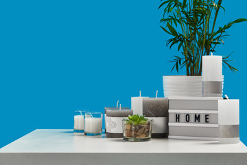 White table with green plant and succulent in pots, some different sized and colors candles, lamp with inscription home. Blue background. Close up