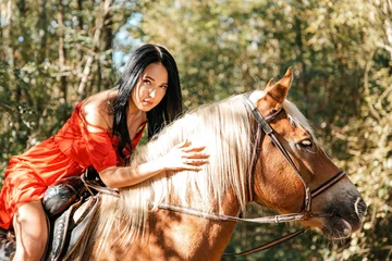 Foto op Canvas A young beautiful brunette woman in a red dress sits astride a brown horse. Summer. Sunlight © _KUBE_