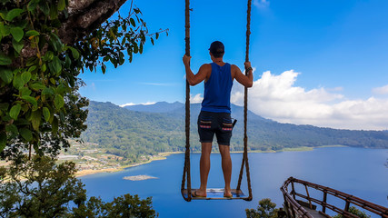 Man enjoying his time on the swing with The Twin Lake behind, Bali, Indonesia. The swing is attached to a tree very high above the lake. He is contemplating the view, having fun. Touristic attraction