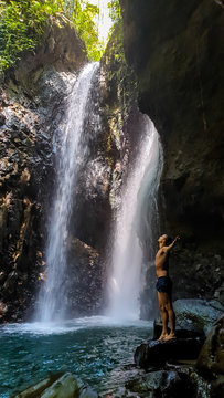 A man in swimsuit standing on the slippery rocks in front of Twin Waterfall, Bali, Indonesia. The waterfall is surrounded by rock from each side. The man enjoyins the beauty of the nature. Adventure