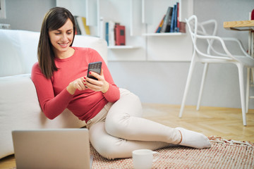 Young smiling woman sitting at home on the floor and using smart phone.