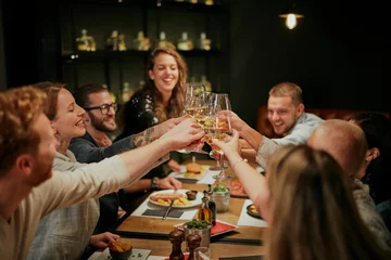 Zelfklevend Fotobehang Beste vrienden die in restaurant zitten voor het diner en een toast maken met witte wijn. Op tafel staat eten. © chika_milan