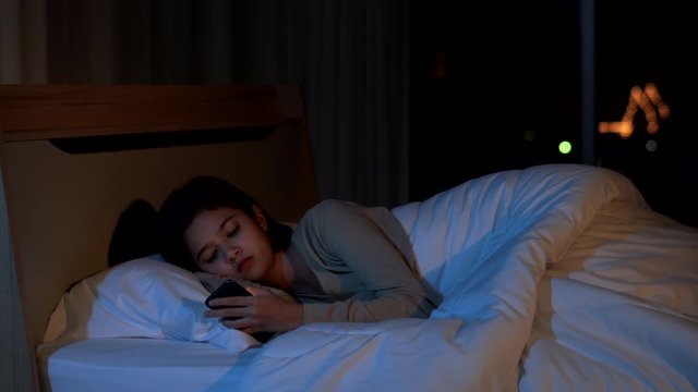 4K Depressed young beautiful Asian woman lying down on the bed and using smartphone. Insomnia sad woman looking at her smartphone. Smartphone addiction, depression symptoms and mental health concept.
