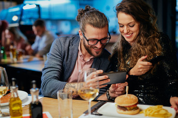 Young man holding smart phone and showing something funny to his grilfriend while sitting in restaurant.